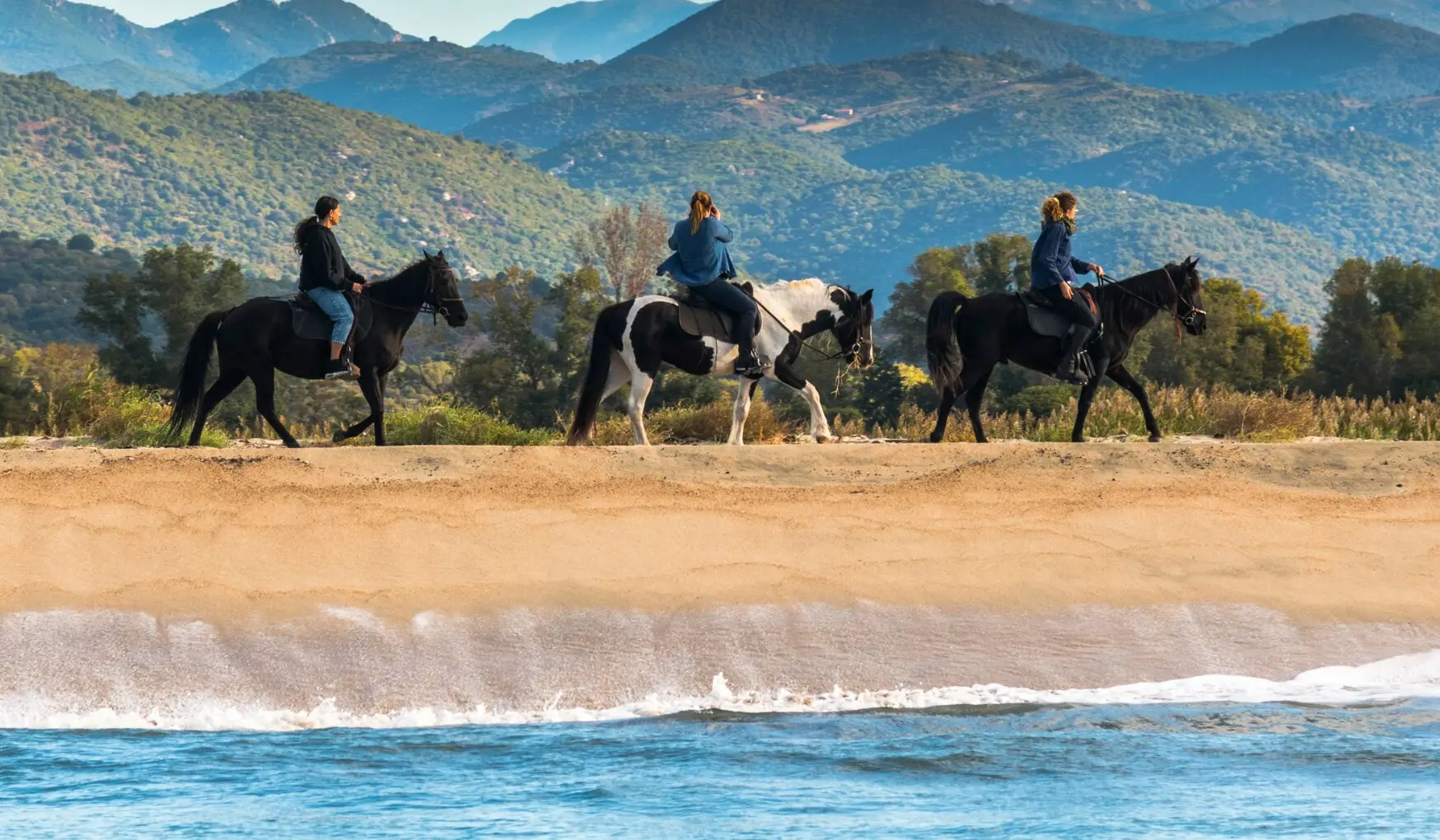 Three riders on L'Orca's horses along the beach at Porto-Pollo, with the Corsican mountains behind
